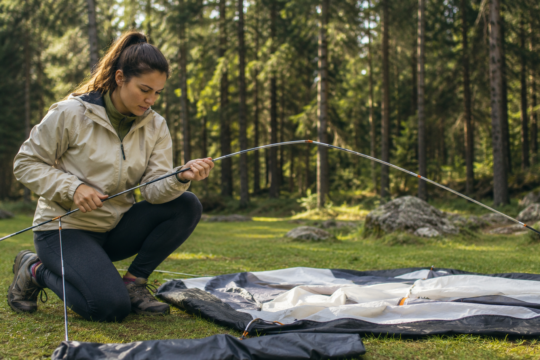 how to pitch a tent camper assembling tent poles at campsite in forest sunny afternoon with tent body spread on ground